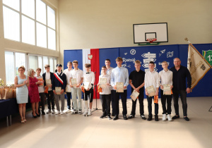 Grupa kilkunastu uczniów i nauczycieli pozuje do wspólnego zdjęcia w sali gimnastycznej. Uczniowie trzymają dyplomy i torby z nagrodami. Po lewej stronie widoczne są wychowawczynie i dyrektorka szkoły, a po prawej nauczyciel w ciemnej koszuli oraz sztandar szkoły. W tle niebieska dekoracja z drogowskazem i godłem Polski.