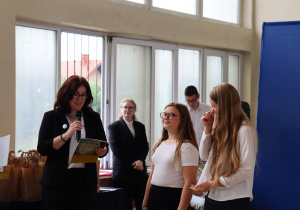 Nauczycielka w ciemnym stroju, trzymając mikrofon i teczkę, przemawia do dwóch uczennic ubranych na biało. Jedna z nich z uśmiechem słucha, druga wzruszona ociera łzy. W tle widać inne osoby w eleganckim stroju i rząd papierowych toreb z upominkami.