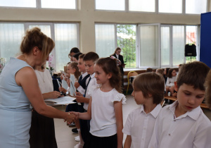 Nauczycielka w jasnoniebieskiej sukience ściska dłoń uśmiechniętej dziewczynki w białej bluzce. Obok stoją uczniowie w galowych strojach, trzymając świadectwa. W tle widać inne dzieci, nauczycieli oraz stoliki z dokumentami i kwiatami.