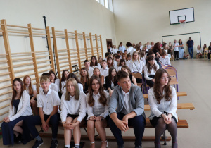 Zdjęcie przedstawia uczniów klas ósmych siedzących na ławkach w sali gimnastycznej podczas uroczystości zakończenia roku szkolnego. Wszyscy są odświętnie ubrani, patrzą w stronę sceny. W tle widać nauczycieli, rodziców i uczniów młodszych klas. Sala udekorowana jest skromnie – bezpośrednio widoczne są przyrządy gimnastyczne i kosz do koszykówki.