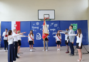 Na zdjęciu uczniowie biorą udział w uroczystości przekazania sztandaru. Pośrodku stoi poczet sztandarowy z widocznym czerwonym sztandarem szkoły. Po lewej i prawej stronie uczniowie wyciągają prawe dłonie w geście ślubowania. W tle niebieska dekoracja z drogowskazami zawierającymi słowa: „marzenia”, „pasja”, „wiedza”, „przygoda” oraz godło Polski i logo Szkoły Podstawowej nr 13 w Pabianicach.