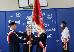 Na zdjęciu uczniowie w galowych strojach z biało-czerwonymi szarfami przekazują sobie czerwony sztandar szkolny. Jeden z uczniów trzyma drzewiec, a drugi go odbiera. W tle dekoracja z napisem „Ruszaj w swoją podróż” i symbolami drogi oraz logo szkoły. Obok stoją inni członkowie pocztu sztandarowego i osoba z mikrofonem.