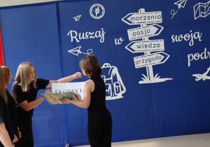 Cztery uczennice w czarnych strojach biorą udział w inscenizacji. Jedna z nich trzyma dużą otwartą książkę, inna wkłada do niej kartkę z napisem „SŁOWO”. W tle dekoracja z hasłem „Ruszaj w swoją podróż” i drogowskazem wskazującym: marzenia, pasja, wiedza, przygoda.