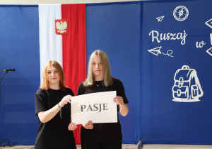Dwie uczennice ubrane na czarno stoją na tle szkolnej dekoracji i trzymają kartkę z napisem „PASJE”. W tle widoczna flaga Polski i fragment dekoracji z napisem „Ruszaj w swoją podróż” oraz grafika drogowskazu.