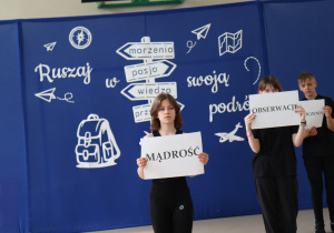 Troje uczniów ubranych na czarno stoi na tle dekoracji z napisem „Ruszaj w swoją podróż” i grafiką drogowskazu. Trzymają kartki z dużymi napisami: „MĄDROŚĆ”, „OBSERWACJE” i częściowo widoczne „DOŚWIADCZENIA”.