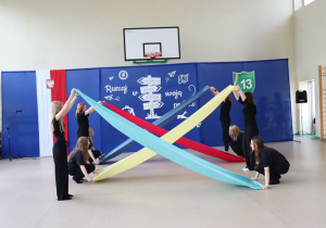 Grupa uczennic ubranych na czarno wykonuje występ artystyczny z użyciem długich, kolorowych szarf (żółtej, niebieskiej, czerwonej i granatowej), tworząc efektowny układ w kształcie krzyża na tle dekoracji z hasłem „Ruszaj w swoją podróż”. W tle widoczny jest kosz do koszykówki i godło Polski.