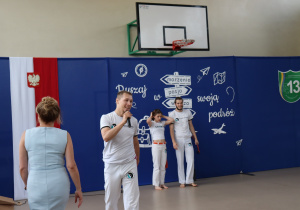 Na zdjęciu instruktor capoeiry przemawia do mikrofonu, a za nim stoją dwie osoby ubrane w białe stroje z emblematem szkoły capoeiry. W tle dekoracja z hasłem „Ruszaj w swoją podróż” i symbolem szkoły nr 13. Po lewej stronie kadru widoczna kobieta w błękitnej sukience.