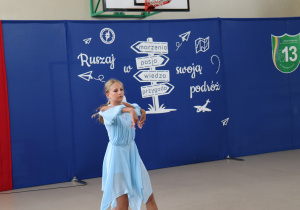 Na zdjęciu dziewczynka w błękitnej sukience wykonuje taniec solowy. Jej ramiona są uniesione w ekspresyjnym geście. W tle dekoracja z niebieskim tłem, napisem „Ruszaj w swoją podróż” oraz drogowskazami z hasłami: „marzenia”, „pasja”, „wiedza”, „przygoda”. Widoczne są też godło Polski i logo Szkoły Podstawowej nr 13 w Pabianicach.