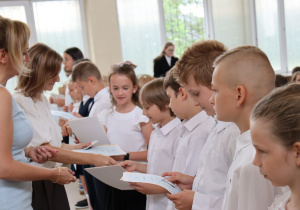 Nauczycielka w jasnoniebieskiej sukience ściska dłoń uśmiechniętej dziewczynki w białej bluzce. Obok stoją uczniowie w galowych strojach, trzymając świadectwa. W tle widać inne dzieci, nauczycieli oraz stoliki z dokumentami i kwiatami.