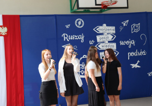 Pięć uczennic w białych i czarnych strojach wykonuje piosenkę, trzymając mikrofony. Stoją na tle dekoracji z hasłem „Ruszaj w swoją podróż” oraz symbolem drogowskazu. W tle widoczna jest flaga Polski i tablica szkolna z numerem 13.