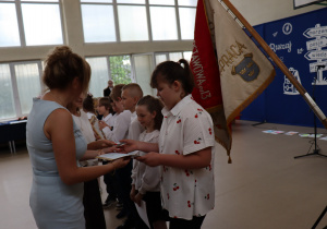 Zdjęcie przedstawia uczniów klas trzecich ubranych na galowo, którzy odbierają świadectwa z rąk nauczycielek podczas uroczystości zakończenia roku szkolnego. W tle widoczny jest sztandar szkoły oraz dekoracja z hasłem „Ruszaj w swoją podróż”.