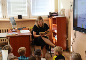 Nauczycielka Anna Wlazeł siedzi w sali lekcyjnej i czyta książkę grupie dzieci siedzących przed nią. W tle znajduje się komputer, tablica interaktywna i półki z pomocami naukowymi.