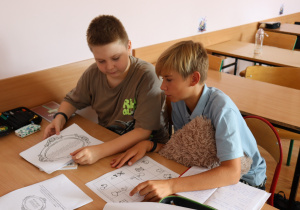 Dwóch uczniów klasy V siedzi przy szkolnej ławce i wspólnie analizuje materiały edukacyjne. Na stoliku znajdują się wydruki z grafikami i tekstem, piórniki oraz zeszyty. Jeden z chłopców przytula pluszową poduszkę, obaj są skoncentrowani na zadaniu.