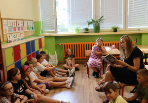 Pani Katarzyna Szczepaniak czyta książkę dzieciom w klasie. Nauczycielka siedzi na krześle, a dzieci uważnie jej słuchają – niektóre siedzą na podłodze, inne na krzesłach. W tle widać kolorowe szafki, plansze edukacyjne oraz rośliny na parapecie. Panuje ciepła i skupiona atmosfera.