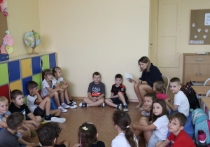 Pani Agnieszka Jarzębowska czyta uczniom klasy pierwszej siedzącym w kręgu na podłodze. Dzieci uważnie słuchają nauczycielki. W tle kolorowe szafki, globus, tablice z dekoracjami oraz biurko z materiałami. Atmosfera skupienia i wspólnej pracy.