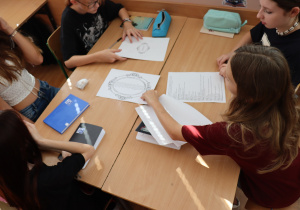 Grupa uczniów klasy V pracuje wspólnie przy jednym stole. Na blacie leżą karty pracy z ćwiczeniami literackimi oraz zeszyty i piórniki. Uczniowie są zaangażowani i skupieni na wykonywanym zadaniu. Widać różnorodne materiały edukacyjne – m.in. arkusze z ozdobnymi ramkami oraz listami wyrazów.