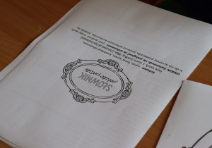 Zbliżenie na kartę pracy zatytułowaną „Słownik podręczny”. Na arkuszu widoczne są ozdobne ramki oraz instrukcja w języku polskim, w której uczniowie proszeni są o wpisanie trudnych słów wraz z definicjami. Kartka leży na drewnianym blacie szkolnej ławki. Widoczna część tekstu zawiera m.in. hasła: „Adrian – wyraz, którym wyrażasz...”, „UWAGA! Rekomendujemy nie posługiwać się przekleństwami...”.