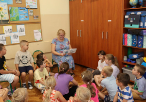 Pani Jolanta Gałkiewicz siedzi na krześle i czyta książkę grupie dzieci, które uważnie jej słuchają. Uczniowie siedzą na podłodze i krzesłach w klasie, której przestrzeń wypełniają kolorowe materiały dydaktyczne, plansze edukacyjne i szafki z pomocami naukowymi. W tle widoczne są plakaty z zasadami zachowania i mapami.