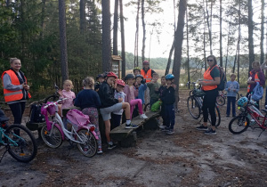 Grupa uczniów klasy 2b z opiekunami podczas rowerowej wycieczki leśnej. Dzieci w kaskach i kamizelkach odblaskowych odpoczywają na polanie wśród drzew.