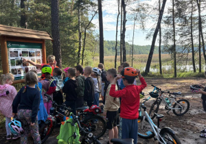 Dzieci z klasy 2b oglądają leśną tablicę edukacyjną o ochronie lasu podczas wycieczki rowerowej z wychowawcami, w tle widoczne drzewa i jezioro.