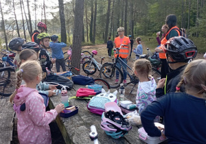 Dzieci z klasy 2b jedzą śniadanie przy stole podczas wycieczki rowerowej. W tle wychowawczyni p. Monika Mettler oraz rodzice w kamizelkach i zaparkowane rowery.