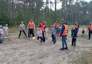 Dzieci z klasy 2b grają w piłkę z wychowawczynią i opiekunami na leśnej polanie. Wszyscy uczestnicy noszą odblaskowe kamizelki i świetnie się bawią.