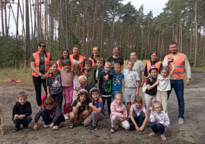 Zdjęcie grupowe uczniów klasy 2b z wychowawczynią Moniką Mettler i rodzicami na tle lasu. Dzieci w kaskach rowerowych, opiekunowie w kamizelkach odblaskowych.