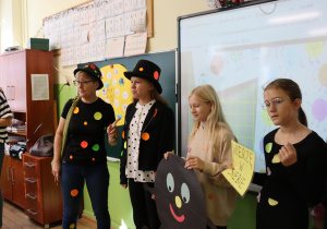 Nauczycielka Monika Mettler i uczennice podczas Dnia Kropki – kolorowe kropki na ubraniach, uśmiechnięta kartonowa postać i hasło „Wierzę w siebie” w klasowej scenerii.