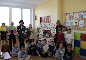 Klasa 1b z wychowawczynią Agnieszką Jarzębowską podczas Dnia Kropki. Uczniowie pozują do wspólnego zdjęcia z kolorowymi kropkami na ubraniach. W tle widać dekoracje z pracami plastycznymi i plakatem „Dzień Kropki”.