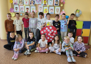 Uczniowie klasy 1b Szkoły Podstawowej nr 13 w Pabianicach pozują wspólnie podczas obchodów Dnia Kropki. Dzieci ubrane są w stroje z kolorowymi kropkami. Na środku znajduje się czerwony plakat – „Kropka Mocy” z rysunkami i hasłami. W tle widać galerię portretów i kolorową dekorację.