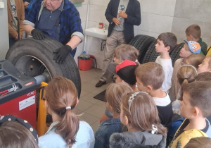 Uczniowie SP 13 słuchają wulkanizatora prezentującego maszynę do wyważania opon. Dzieci z zaciekawieniem obserwują pokaz, wokół stoją nauczyciele i pracownicy MZK.