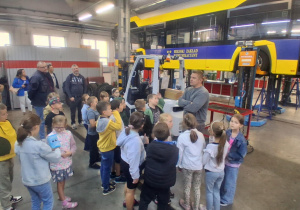 Uczniowie klasy 2B w hali MZK słuchają pracownika opowiadającego o naprawie autobusów. W tle uniesiony pojazd serwisowy, dzieci stoją w półkolu i zadają pytania.