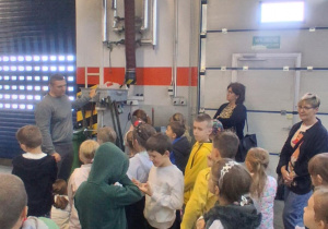 Uczniowie klasy 2B słuchają pracownika MZK, który prezentuje urządzenie warsztatowe. W tle nauczycielki Monika Juran-Tłoczek i Monika Mettler obserwują zajęcia.