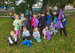 Grupa dzieci z kolorowymi workami i rękawiczkami po zakończonej akcji „Sprzątanie Świata” stoi pod drzewami, prezentując zebrane śmieci.