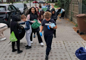 Grupa uczniów SP13 zbierająca śmieci przy ulicy podczas akcji Sprzątania Świata; niosą worki, mają rękawiczki ochronne, w tle drzewa i samochody.