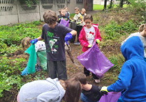 Uczniowie sprzątają teren wokół szkoły podczas akcji „Sprzątanie Świata” – dzieci z kolorowymi workami zbierają śmieci w zarośniętym miejscu przy ogrodzeniu.