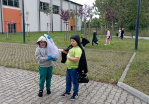 Dwóch uczniów SP13 prezentuje worek śmieci podczas akcji Sprzątanie Świata, w tle uczniowie porządkują teren zielony przy szkole.