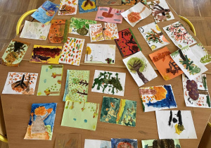 Na stole leżą jesienne prace plastyczne dzieci — kolorowe obrazki z liśćmi, drzewami i krajobrazami wykonane farbami, kredkami i innymi technikami.