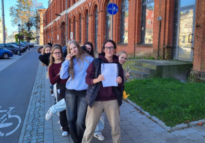 Pani Agnieszka Skowron z uczennicami klasy 8a podczas spaceru edukacyjnego śladami niemieckiego dziedzictwa w Pabianicach, stoją przed ceglanym budynkiem fabrycznym.
