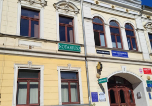 Zabytkowa kamienica w Pabianicach z rzeźbami na fasadzie i szyldem "Notariusz". Przed budynkiem rozkwitłe różowe krzewy, na tle niebieskiego nieba.