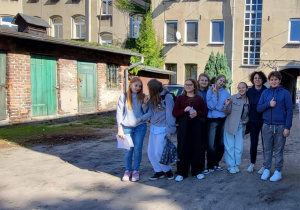 Pani Sylwia Muszyńska i uczniowie klasy 8a stoją na podwórku przy kamienicy z zielonymi drzwiami komórek. W tle jasna elewacja i słońce rzucające cienie drzew.