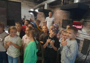Grupa dzieci przy piecu chlebowym z rozżarzonym wnętrzem, w tle piekarz opowiadający o wypieku chleba.