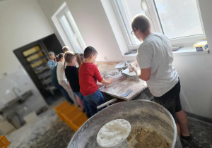 Dzieci uczą się wyrabiać ciasto w piekarni, przy stole pod oknem. Piekarz pokazuje technikę pracy. Na pierwszym planie misa z ciastem.