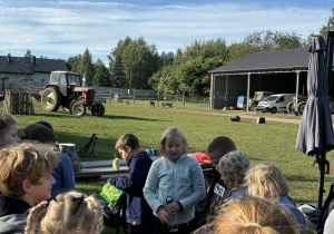 Dzieci uczestniczą w zajęciach na świeżym powietrzu na wiejskim podwórzu. W tle traktor, budynki gospodarcze i łąka.
