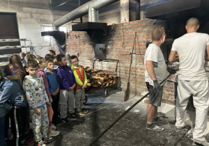 Uczniowie klasy 2A i 2B obserwują piekarzy wkładających chleb do pieca opalanego drewnem wewnątrz tradycyjnej piekarni.