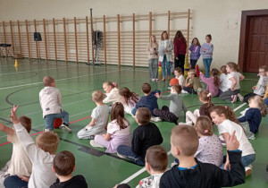 Dzieci siedzące na sali gimnastycznej podczas Dnia Integracji, prowadzące z mikrofonami i nauczycielka stojące z tyłu.