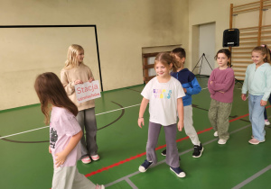 Dziewczynka trzymająca tabliczkę „Stacja uśmiechów” podczas zabawy integracyjnej; uczniowie przechodzą obok niej z uśmiechami – scena z Dnia Integracji w sali gimnastycznej.