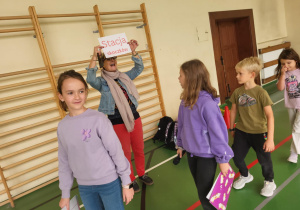 Dzieci przechodzą obok nauczycielki z tabliczką „Stacja skoczków” podczas szkolnej gry integracyjnej na sali gimnastycznej.