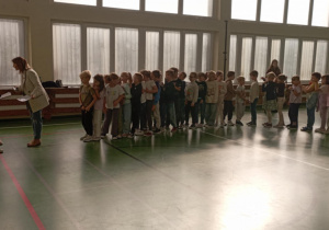 zieci stojące w dwóch rzędach na sali gimnastycznej tworzą „Pociąg przyjaźni”; z przodu uczennice i nauczycielka koordynują przebieg wydarzenia.