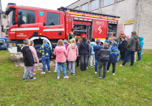 Grupa dzieci z klasy II stoi przy wozie strażackim OSP Pabianice, słuchając dwóch strażaków prezentujących wyposażenie ratownicze.